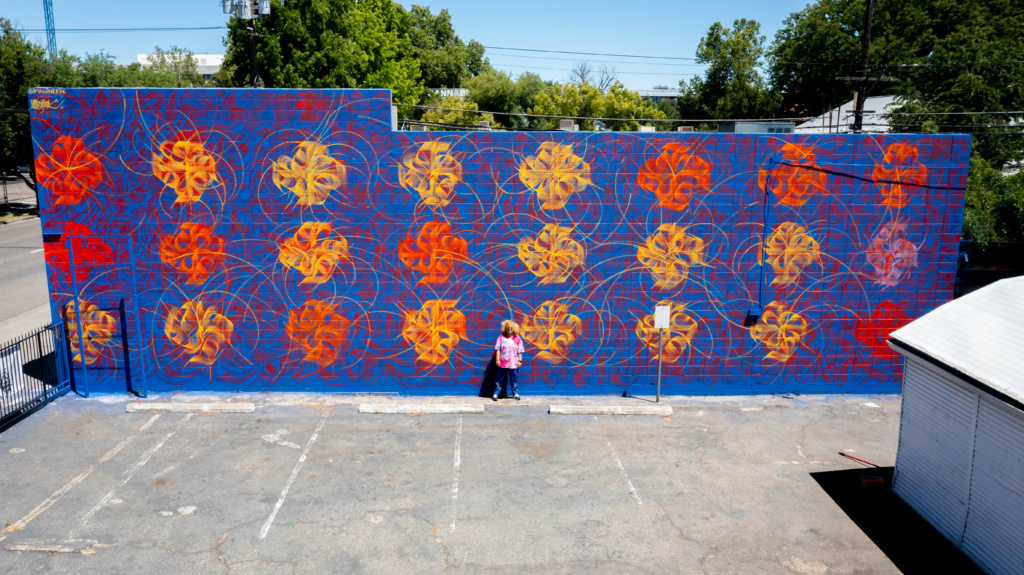 Artist Imagine stands in front of her mural for the Wide Open Walls Festival.  The mural features red and yellow pinwheel-like images on a blue background.